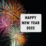 Black "Happy New Year 2022" text on a white square background. White background is on right side of an image of orange, green, and purple fireworks in a dark night time sky. 