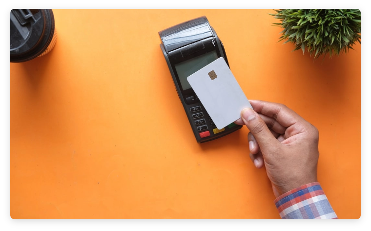 A hand taps a credit card on a card reader on a bright orange table