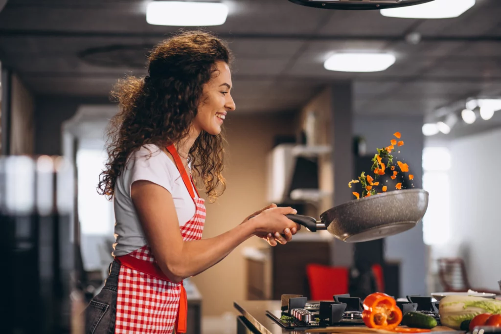 Woman leading cooking class. She’s wearing an apron and flipping vegetables in a heated pan.