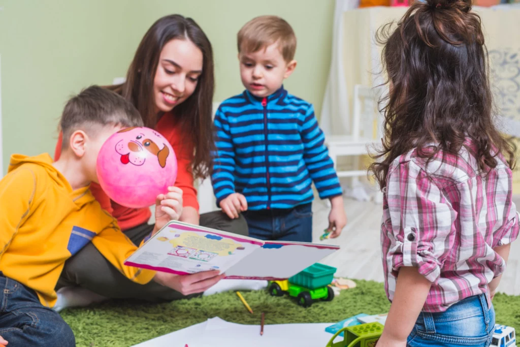 Female nanny reading to three young children. They’re sitting on the floor. The children are playing with toys.