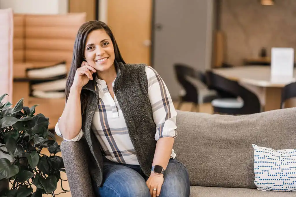 Photo of professional career coach, Angie Callen. She is sitting on a gray couch. She has brown hair and is wearing a gray vest over a white and brown shirt