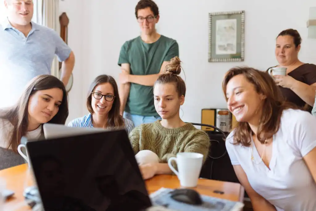 4 coworkers sit together around a wooden table, looking at a laptop.