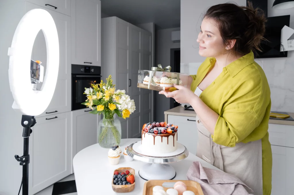 Social media influencer in yellow dress and apron using circle light to record on phone. She is standing in the kitchen, behind a counter with cake and several bakery and pastry items. In her hands, she’s showing off a gift box of cupcakes.