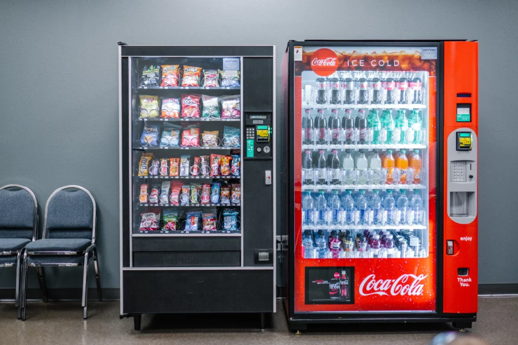 Two vending machines stand side by side inside an office. One sells Coca-Cola drink products. The other vending machine sells chips, candy, and other miscellaneous snacks. Both machines accept cash and have a credit card reader.