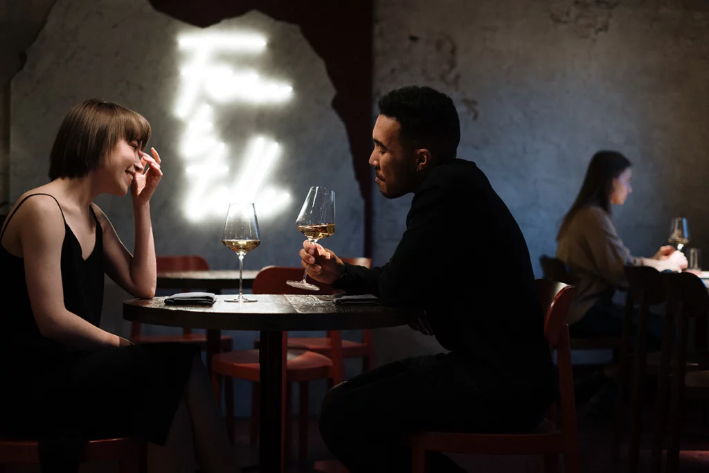 A couple enjoys a romantic date night at a dimly-lit restaurant. Each table seems to have its own separate lighting fixture. The couple chats while holding wine glasses.